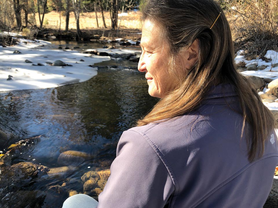 photo of Deborah Goslin sitting by a river in winter, absorbing sunlight with her eyes closed.