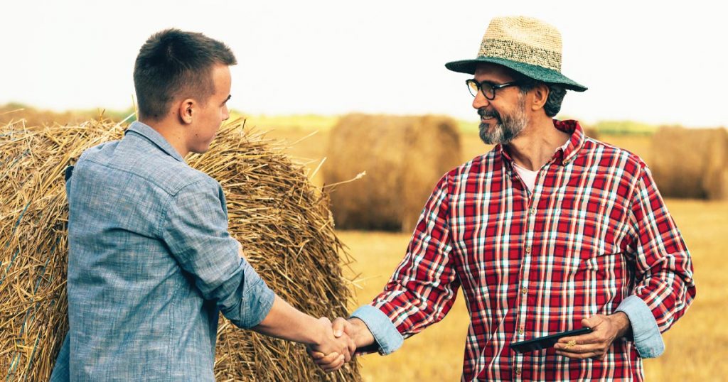 Photo of two men shaking hands in a field with round hay bails.